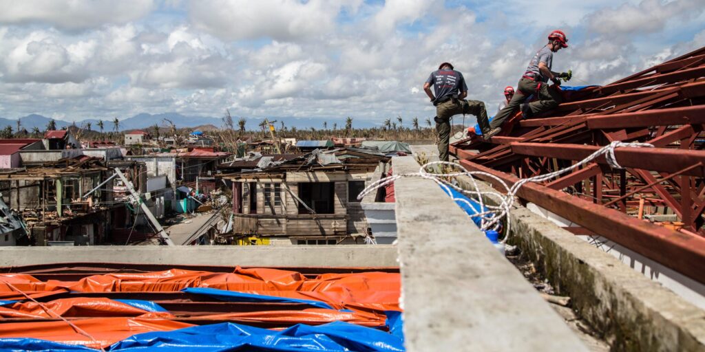 Volunteers stand on roof beams to tarp a roof with homes highly damaged by Typhoon Haiyan in the background. 
