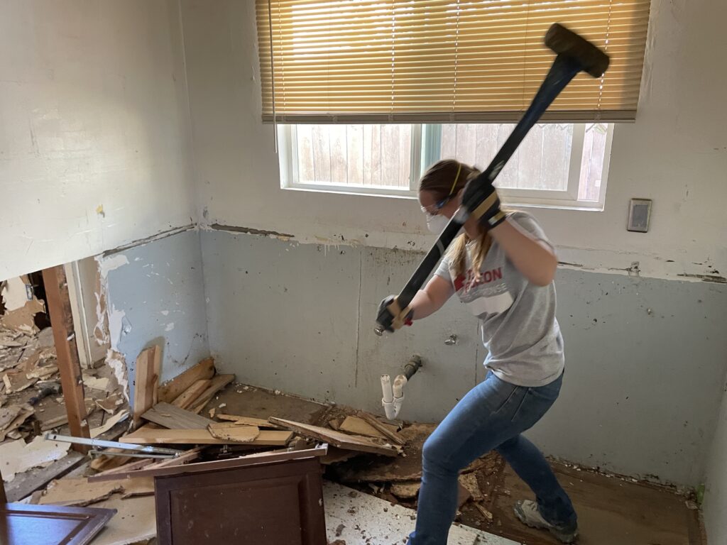 Female volunteer demolishes wall with sledgehammer.